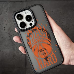 Puede incluir: Una funda de smartphone negra con un gráfico de un balón de baloncesto naranja envuelto en llamas naranjas. El texto "PLAY HARD" está impreso en naranja, con las palabras "STAY TRUE" centradas en el balón. La funda tiene un acabado mate.