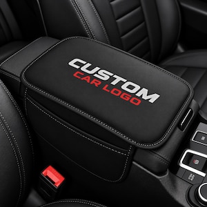 May include: A black car center console armrest cover featuring the white text CUSTOM and the red text CAR LOGO printed on the top surface. The cover has visible white stitching along the edges and is installed on a vehicle center console.