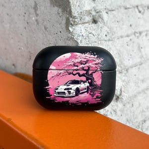 Könnte beinhalten: Schwarzes Schutzetui für Ohrhörer mit einer Grafik eines weißen Sportwagens unter einem rosa Kirschblütenbaum und einer rosa Sonne. Das Etui liegt auf einer orangefarbenen Oberfläche.