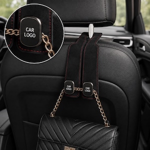 Puede incluir: Un gancho de ante negro para el reposacabezas del coche con costuras rojas y un emblema metálico con el texto CAR LOGO. El gancho incluye una cadena dorada diseñada para sujetar un bolso negro acolchado contra el respaldo del asiento.