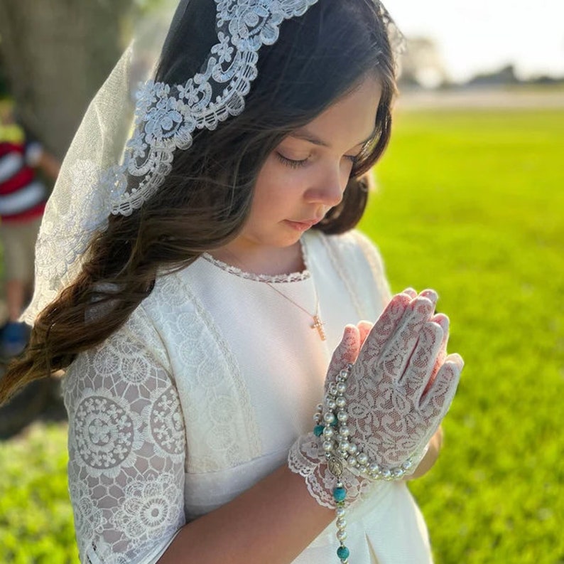 May include: A young girl wearing a white lace dress and a white lace veil. She is holding her hands together in prayer and wearing a white lace glove on one hand. She is also wearing a necklace with a cross pendant.