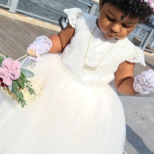 May include: A young girl in a white lace dress and white gloves holds a flower basket with pink flowers and greenery. She is wearing a gold necklace with a nameplate pendant.