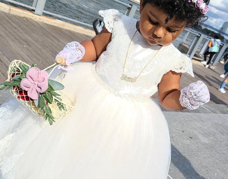 May include: A young girl in a white lace dress and white gloves holds a flower basket with pink flowers and greenery. She is wearing a gold necklace with a nameplate pendant.