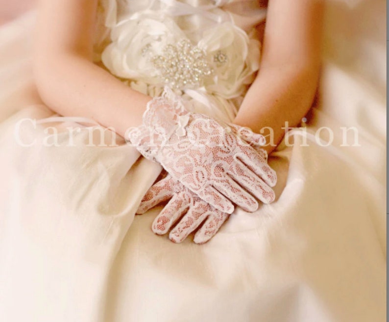 May include: A pair of white lace gloves with a floral pattern. The gloves are worn by a person wearing a white dress.