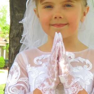 May include: A young girl wearing a white lace dress and veil, with her hands clasped in prayer.