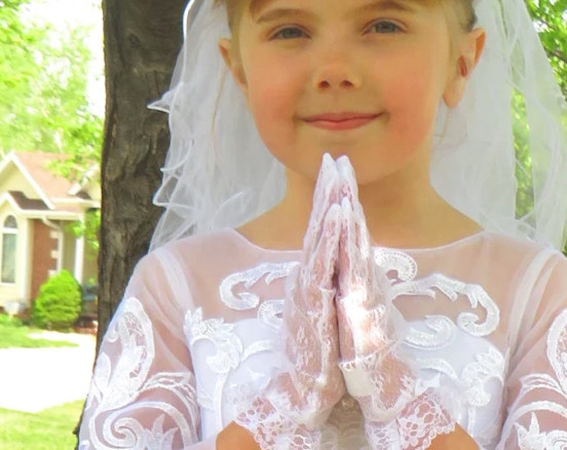 May include: A young girl wearing a white lace dress and veil, with her hands clasped in prayer.