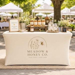 May include: A cream-colored fitted tablecloth featuring a logo with a bee and wildflowers above the text Meadow & Honey Co. Local Honey Pure & Raw. The table displays jars of honey, small bouquets of white flowers, and two signs with text.