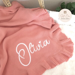 May include: A dusty rose knitted blanket featuring a ruffled border and the name Olivia embroidered in white script with two small heart accents above the letters. A circular tag in the corner displays the text Gift Box AVAILABLE.