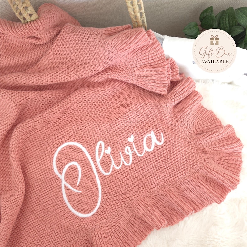 May include: A dusty rose knitted blanket featuring a ruffled border and the name Olivia embroidered in white script with two small heart accents above the letters. A circular tag in the corner displays the text Gift Box AVAILABLE.