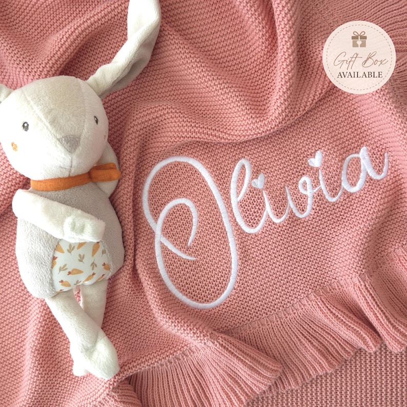 May include: A dusty pink knitted blanket with a ruffled edge and the name Olivia embroidered in white script. A plush rabbit toy with long ears, an orange scarf, and a carrot-patterned belly sits on the blanket. A circular tag reads Gift Box Available.