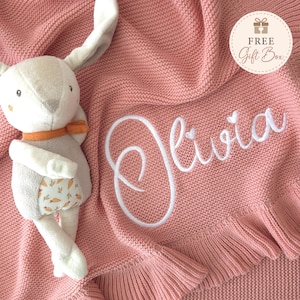 May include: A dusty pink knitted blanket with a ruffled edge and the name Olivia embroidered in white script. A plush rabbit toy with long ears, an orange scarf, and a carrot-patterned belly sits on the blanket. A circular tag reads FREE Gift Box.