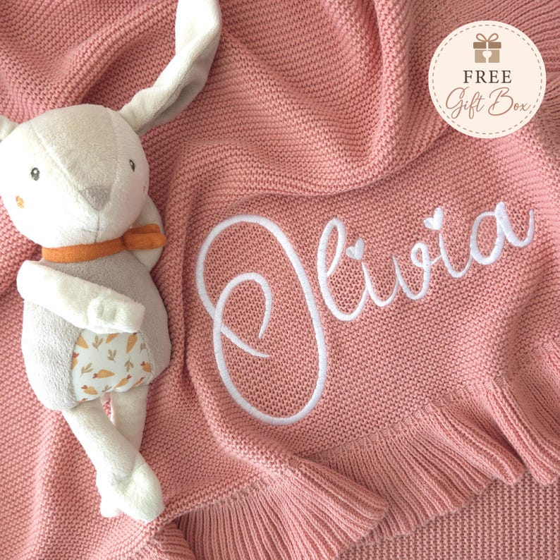 May include: A dusty pink knitted blanket with a ruffled edge and the name Olivia embroidered in white script. A plush rabbit toy with long ears, an orange scarf, and a carrot-patterned belly sits on the blanket. A circular tag reads FREE Gift Box.