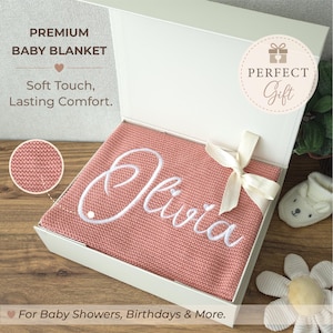 May include: A dusty pink knitted baby blanket with the name Olivia embroidered in white script, presented in a white gift box with a cream ribbon. Text on the box reads PREMIUM BABY BLANKET, Soft Touch, Lasting Comfort, and PERFECT Gift.