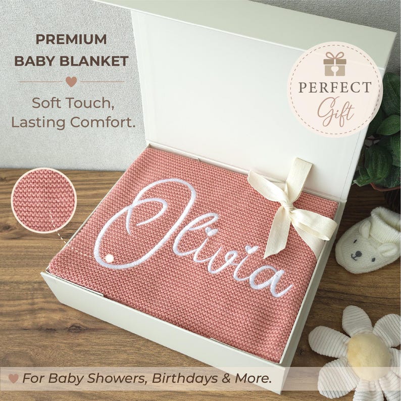 May include: A dusty pink knitted baby blanket with the name Olivia embroidered in white script, presented in a white gift box with a cream ribbon. Text on the box reads PREMIUM BABY BLANKET, Soft Touch, Lasting Comfort, and PERFECT Gift.