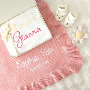 May include: A pink knitted baby blanket with a ruffled edge, embroidered with the white text "Sophia Elise" and "12.05.2026". A white gift box contains a white blanket with the pink embroidered name "Gianna", two white bear booties, and a flower toy.
