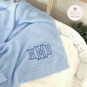 Può includere: Una coperta lavorata a maglia azzurra con una trama testurizzata e un bordo a coste. La coperta è ricamata con le lettere del monogramma blu scuro NMB in un carattere serif decorativo. Un badge bianco circolare mostra il testo Gift Box AVAILABLE.
