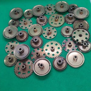 Op de afbeelding: Een verzameling kleine, messingkleurige metalen tandwielen en katrollen in verschillende maten en uitvoeringen. Sommige hebben cirkelvormige vormen, terwijl andere getande randen hebben. De componenten zijn gerangschikt op een groene ondergrond.