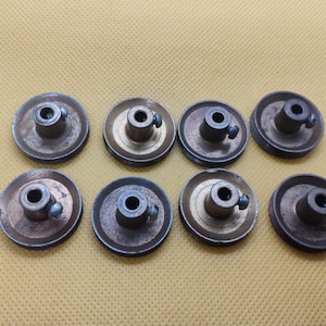 Pode incluir: Oito polias de metal vintage com um cubo central e um pequeno parafuso. As polias têm um design circular com um esquema de cores bronze e prata. Estão dispostas em duas filas de quatro sobre um fundo amarelo.