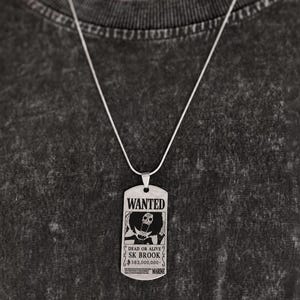 May include: A silver-toned metal dog tag pendant on a thin chain. The pendant features a black engraved graphic of a skeleton character with an afro, the word WANTED, the text DEAD OR ALIVE, SK BROOK, 383,000,000, and the word MARINE at the bottom.