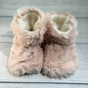 May include: A pair of soft, pink faux fur baby booties. The booties have a fluffy exterior and a cream-colored interior. The design is a cozy, warm style, perfect for keeping little feet comfortable. The booties are set against a light wood-grain background.