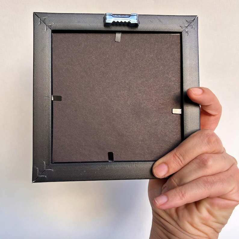 Op de afbeelding: Achterkant van een kleine, vierkante zwarte fotolijst. De lijst heeft een donkerbruine achterkant en metalen clips om de foto op zijn plaats te houden. Een metalen hanger is aan de bovenkant bevestigd. De lijst wordt door een hand vastgehouden.