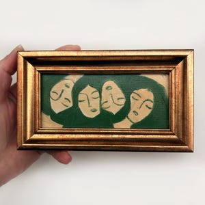 K&ouml;nnte beinhalten: Gerahmtes Gem&auml;lde mit vier stilisierten Gesichtern mit geschlossenen Augen und dunkelgr&uuml;nem Haar vor einem gr&uuml;nen Hintergrund. Das Kunstwerk befindet sich in einem rechteckigen, goldfarbenen Rahmen und erzeugt eine Vintage-&Auml;sthetik.