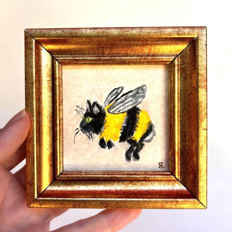 May include: A small, square painting in a gold frame. The artwork depicts a black cat with yellow and black stripes, resembling a bee. The cat has green eyes and wings. The painting is signed in the lower right corner.