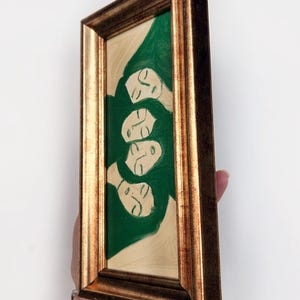 K&ouml;nnte beinhalten: Ein gerahmtes Gem&auml;lde mit vier stilisierten Gesichtern mit geschlossenen Augen, vor einem gr&uuml;nen und beigen Hintergrund. Das Kunstwerk ist in einem goldfarbenen, verzierten Rahmen eingefasst, was dem St&uuml;ck einen Hauch von Eleganz verleiht.