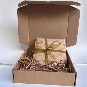 Pu&ograve; includere: Una scatola di cartone marrone aperta, che rivela un pacco avvolto e legato con un nastro verde. Il pacco &egrave; annidato in trucioli di carta marrone. Il pacco ha un'etichetta stampata che dice "THANK YOU SO MUCH".