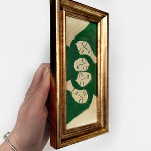 K&ouml;nnte beinhalten: Ein gerahmtes Gem&auml;lde mit einem goldfarbenen Rahmen. Das Kunstwerk zeigt vier stilisierte Gesichter mit geschlossenen Augen, die in Wei&szlig; auf einem gr&uuml;nen Hintergrund gemalt sind. Das Gem&auml;lde ist rechteckig und hat eine vertikale Ausrichtung.