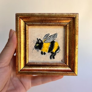 May include: A framed, square painting of a cat with bee wings and yellow and black stripes. The cat has green eyes and whiskers. The frame is gold-colored with a detailed, ornate design.