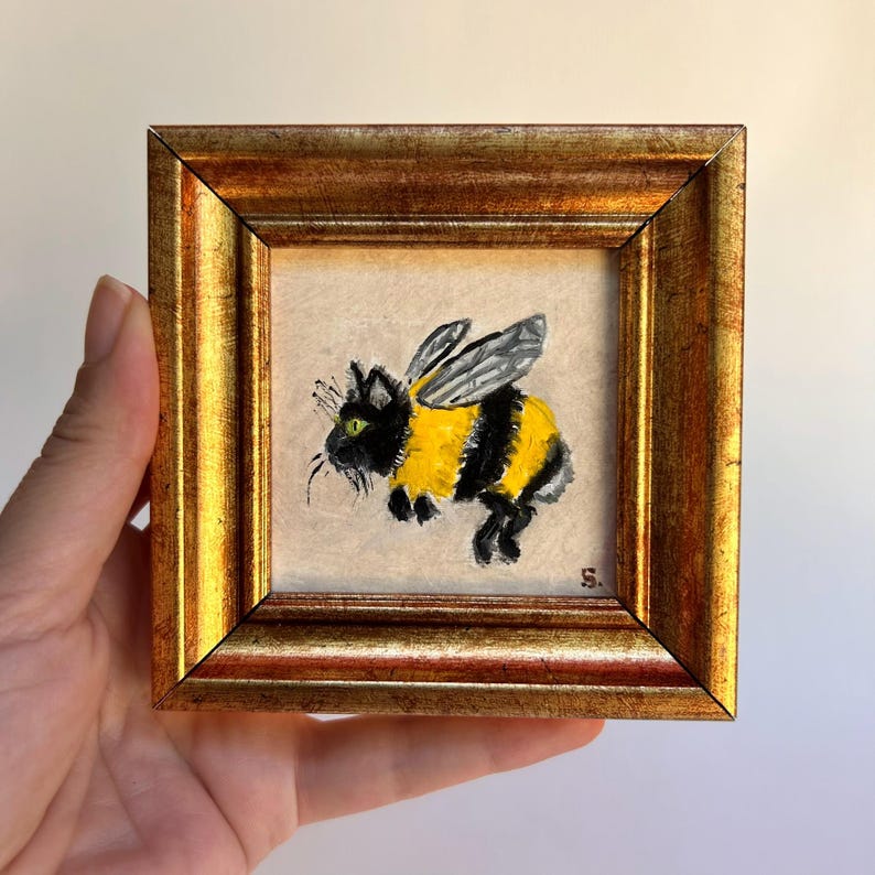May include: A framed, square painting of a cat with bee wings and yellow and black stripes. The cat has green eyes and whiskers. The frame is gold-colored with a detailed, ornate design.