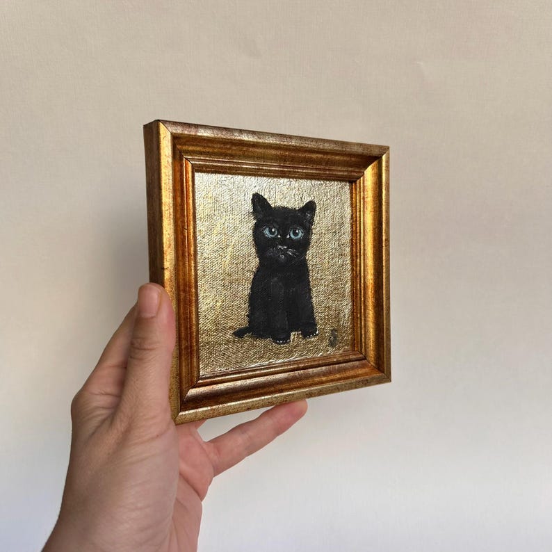 Op de afbeelding: Een kleine, ingelijste schildering van een zwarte kat met helderblauwe ogen. De kat zit rechtop tegen een gouden achtergrond. De lijst is goudkleurig en rechthoekig.