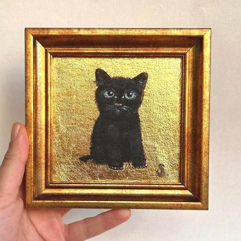 Op de afbeelding: Een kleine, vierkante schildering van een zwarte kat met helderblauwe ogen. De kat is gecentreerd op een gouden, getextureerde achtergrond, omlijst door een goudkleurige houten lijst.