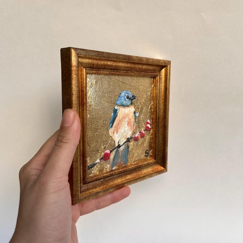 Pu&ograve; includere: Un piccolo dipinto incorniciato di un uccello azzurro appollaiato su un ramo con bacche rosse. L'uccello ha piume blu e pesca. Lo sfondo &egrave; dorato e la cornice &egrave; di un caldo marrone dorato.