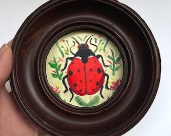 May include: A small, round, framed painting of a ladybug. The ladybug is red with black spots and a black head. The background is a light yellow with green leaves and small pink flowers. The frame is dark brown.