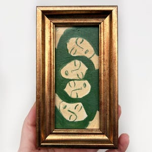 K&ouml;nnte beinhalten: Ein gerahmtes Gem&auml;lde mit einem goldfarbenen Rahmen. Das Kunstwerk zeigt vier stilisierte Gesichter in Creme vor einem dunkelgr&uuml;nen Hintergrund. Das Gem&auml;lde ist rechteckig und wird von einer Hand gehalten.