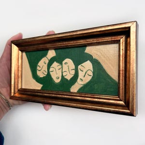 K&ouml;nnte beinhalten: Gerahmtes Gem&auml;lde mit goldfarbenem Rahmen. Das Kunstwerk zeigt vier Gesichter mit geschlossenen Augen, gemalt in Gr&uuml;n- und Beiget&ouml;nen. Das Gem&auml;lde wird von einer Hand gehalten.