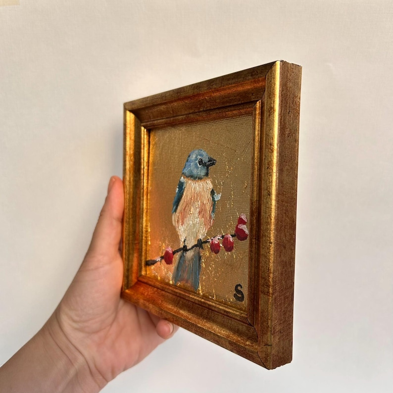 Pu&ograve; includere: Un piccolo dipinto quadrato di un uccello azzurro appollaiato su un ramo con bacche rosse. L'uccello ha piume blu e arancioni. Il dipinto &egrave; in una cornice dorata.