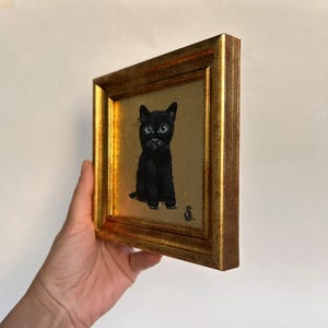 Op de afbeelding: Een ingelijst schilderij van een zwarte kat met helderblauwe ogen. De kat zit rechtop tegen een gouden achtergrond. De lijst is goudkleurig en rechthoekig. De schildering is rechtsonder gesigneerd.