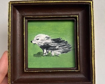 May include: A small, square painting of a hedgehog in a brown frame with a gold inner edge. The hedgehog is depicted with white and black brushstrokes on a green background. The artwork is held by a hand.