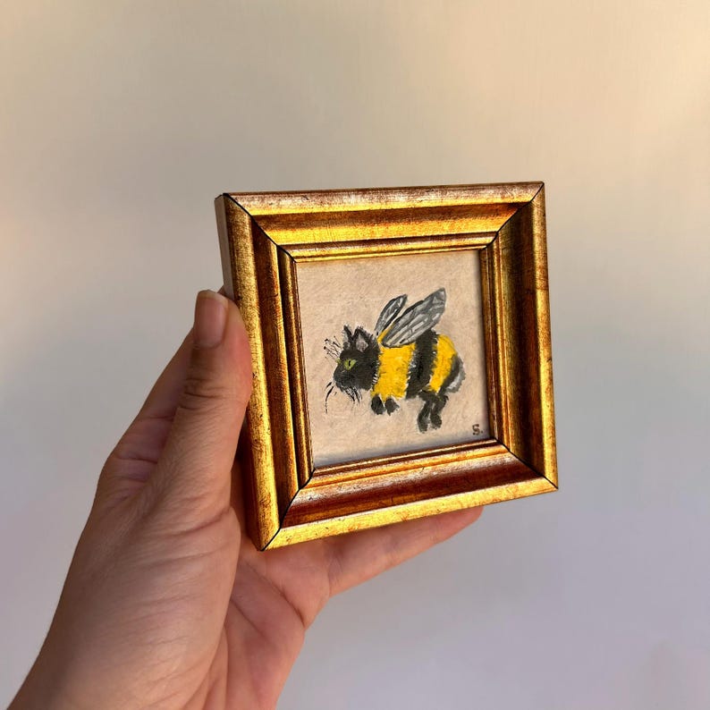 May include: A small, square framed painting featuring a whimsical bee-cat hybrid. The artwork showcases a black and yellow creature with wings, set against a neutral background. The frame is a gold color.