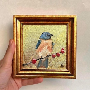 Pu&ograve; includere: Piccolo dipinto quadrato in cornice dorata. L'opera raffigura un'uccello azzurro posato su un ramo con bacche rosse su sfondo dorato. L'uccello ha piume blu e beige. La cornice &egrave; di colore oro caldo.