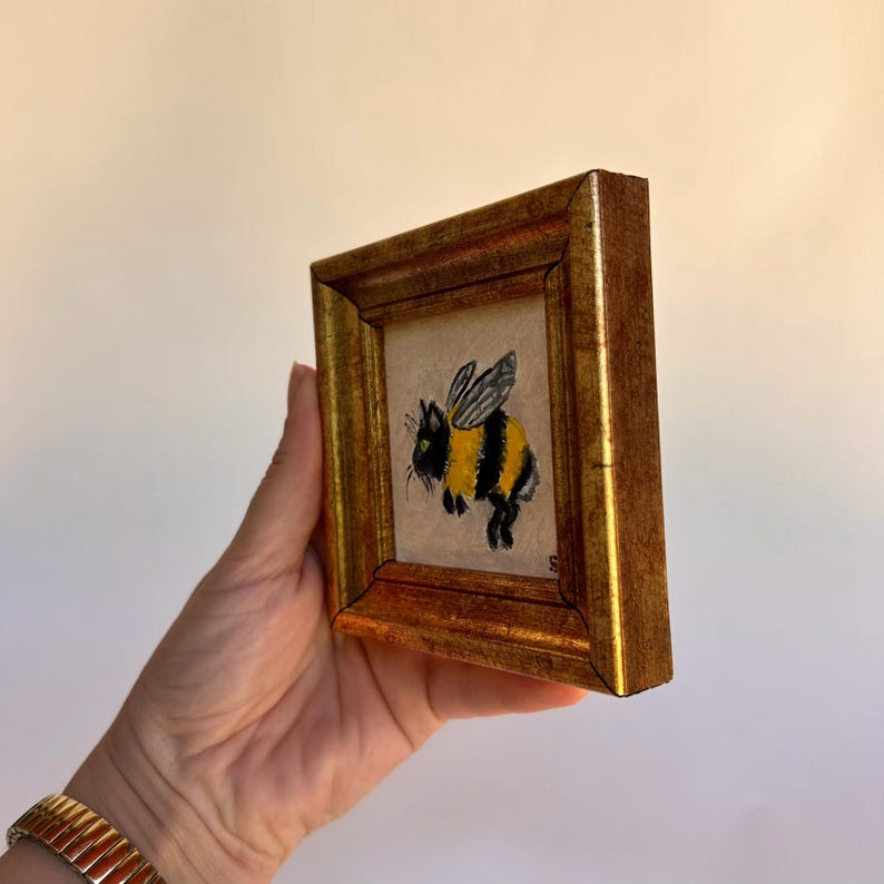 May include: A small framed painting of a bumblebee. The bee is depicted with yellow and black stripes, black legs, and wings. The frame is gold-colored and rectangular.