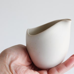 May include: A small, off-white ceramic creamer with a curved spout and a smooth, matte finish. The creamer is held in a hand against a plain white background. The design is minimalist and modern.