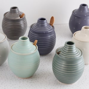 May include: A collection of ceramic honey pots in various colors, including gray, blue, and white. Each pot has a lid and a wooden honey dipper. Some pots have a textured surface with small bumps, while others have horizontal lines.