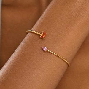 Può includere: Un bracciale aperto color oro con due diverse pietre preziose alle estremità. Un'estremità presenta una pietra arancione rettangolare, mentre l'altra estremità è dotata di una pietra rosa a forma di goccia. Il cinturino in metallo è sottile e lucido.