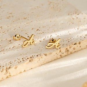 Peut inclure: Une paire de boucles d'oreilles en forme de lettre "A" dorée. Les boucles d'oreilles sont présentées sur une surface texturée blanc cassé. Les boucles d'oreilles sont petites et délicates, avec une police cursive. Le fond est de couleur neutre.