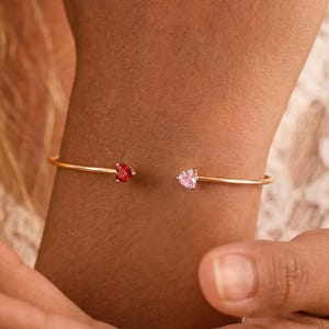 Könnte beinhalten: Ein goldfarbenes, offenes Armband mit zwei herzförmigen Steinen an den Enden. Ein Stein ist tiefrot, der andere zartrosa gefärbt. Das Armband besteht aus einem dünnen, glatten Metallreif, der das Handgelenk mit einer offenen Lücke umschließt.