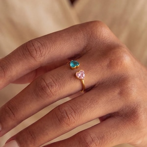 May include: A gold-toned open ring featuring two heart-shaped stones, one in a vibrant blue and the other in a soft pink. The band is thin and metallic, designed to sit comfortably on the finger with the two coloured hearts facing each other.